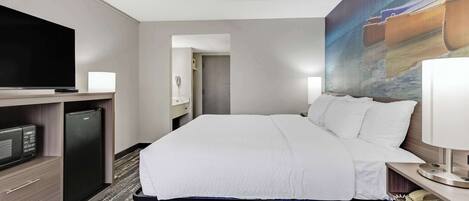 Quarto de hotel com cama, televisão, frigorífico, secretária com candeeiro e um quadro na parede.