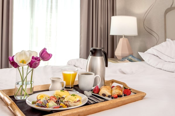 A breakfast tray with eggs benedict, croissants, strawberries, and a coffee pot on a bed with a lamp and curtains in the background.