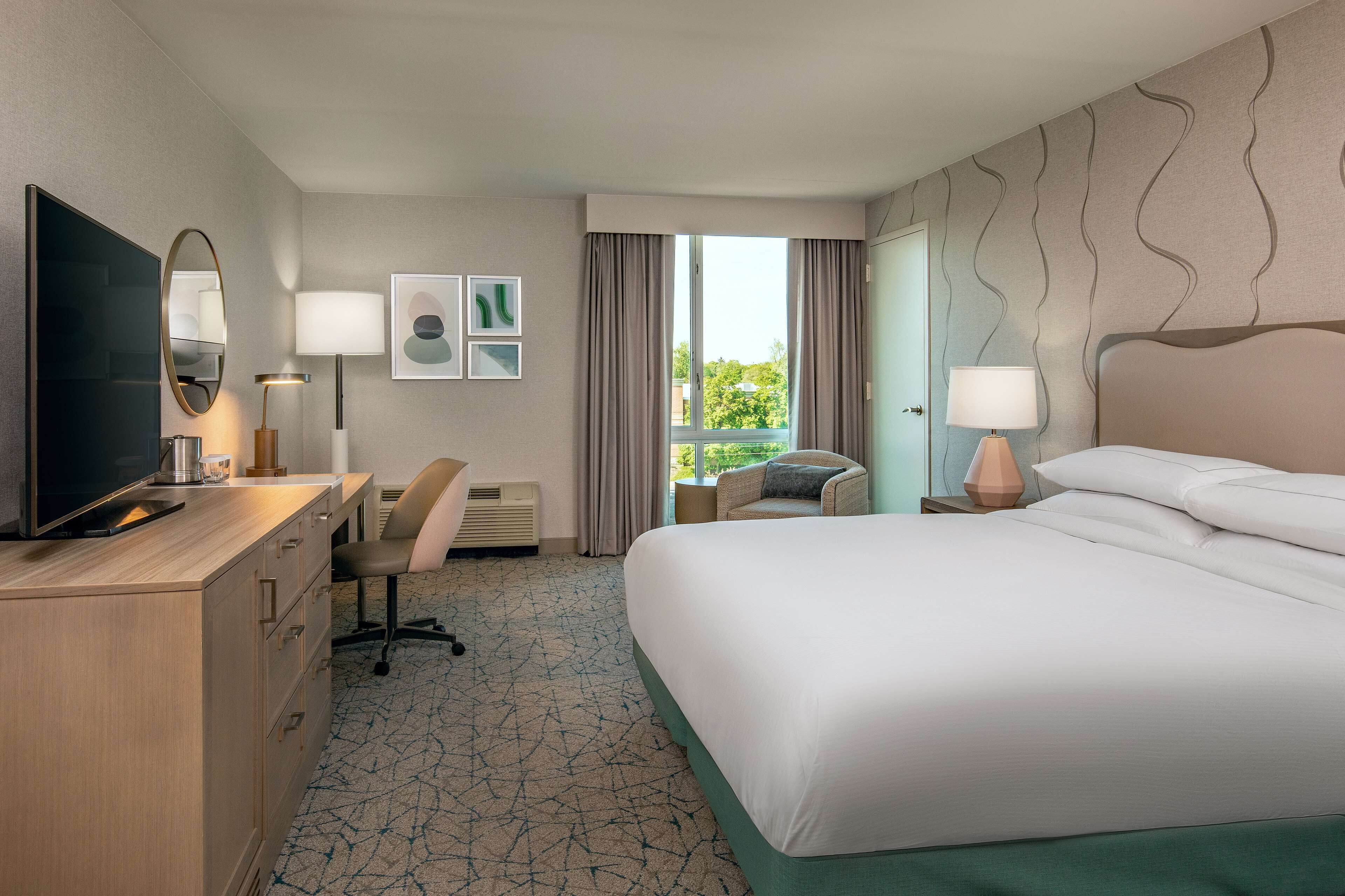 Habitación de hotel con cama, escritorio con televisor, una silla, lámpara y vistas a zonas verdes.