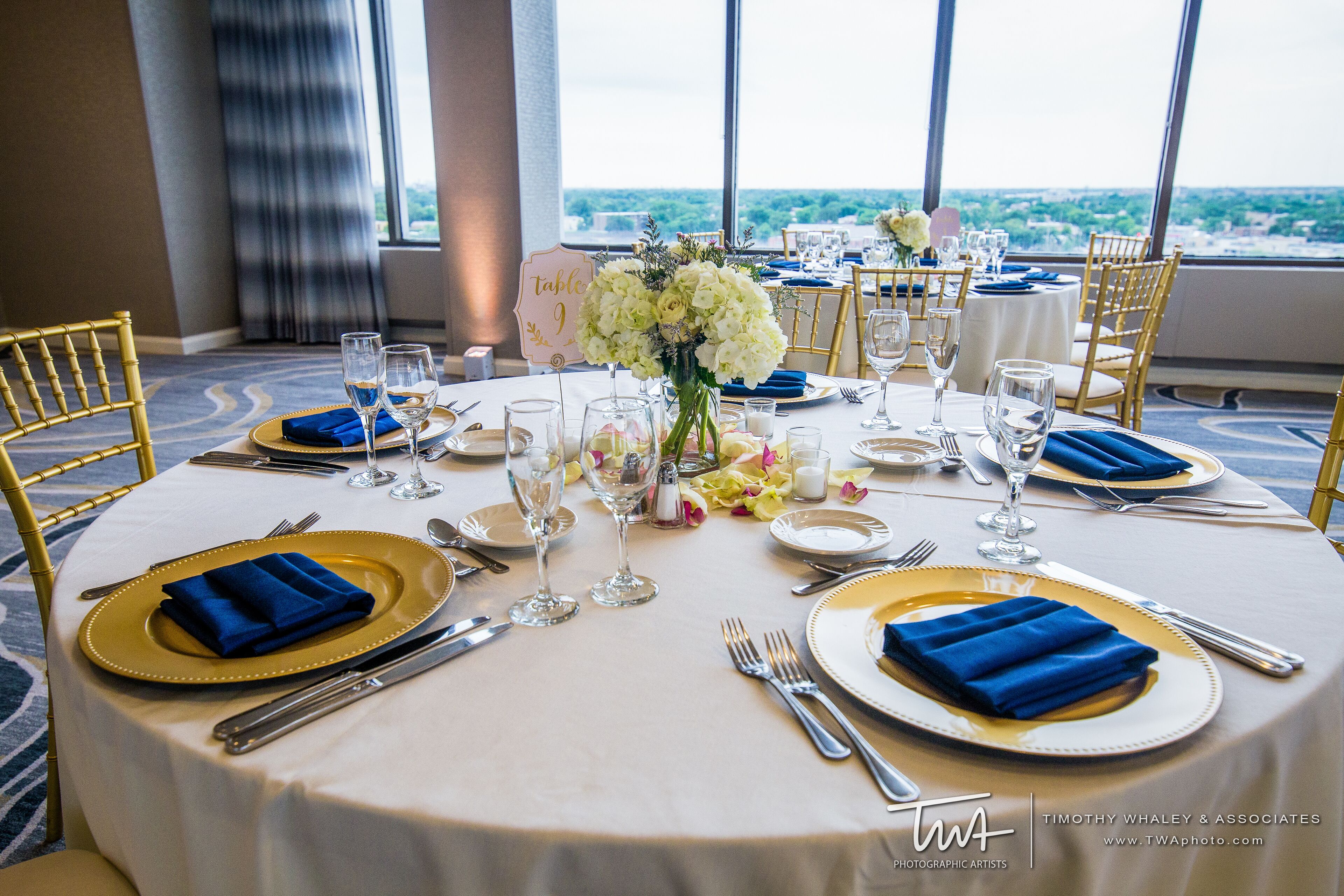 A table set for a formal dinner with gold and blue tableware, white linens, and a view of the city.