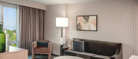 A hotel room with a sofa, armchair, coffee table, lamp, and artwork on the wall.