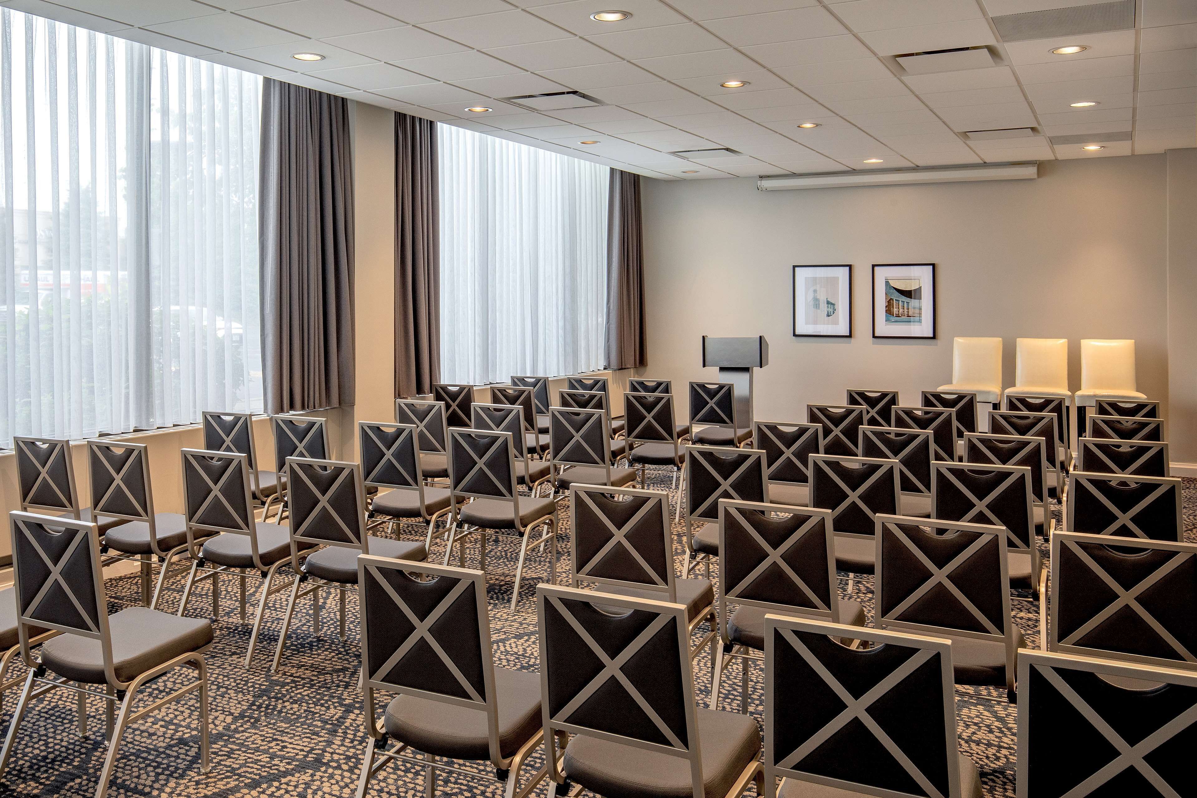 A conference room with rows of chairs, large windows with curtains, and two wall-mounted lamps.