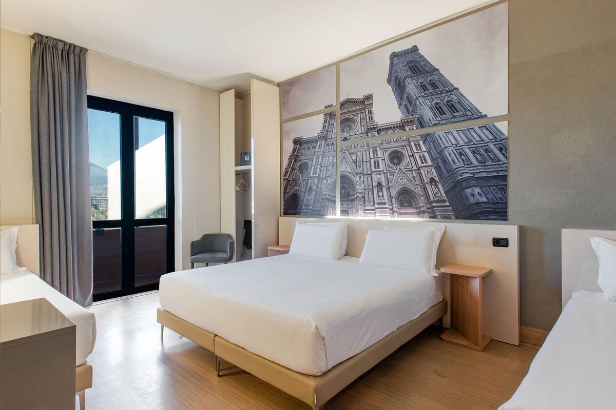 Ein Hotelzimmer mit einem großen Bett, einem Schreibtisch, einem Sessel, einem Nachttisch, einem Bild eines Gebäudes und Blick durch ein Fenster.