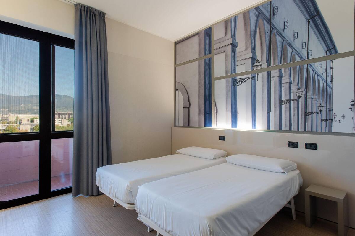 Ein Hotelzimmer mit zwei Betten, einem großen Fenster mit Blick, einem Bild an der Wand und einem kleinen Nachttisch.