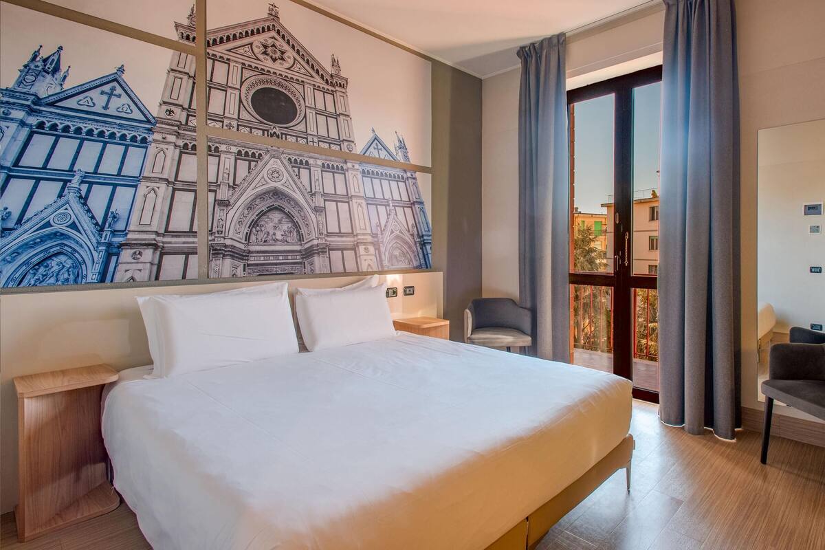 Ein Hotelzimmer mit einem großen Bett, einem Nachttisch, einem Sessel, einem großen Fenster mit Vorhängen und einem Wandbild eines Gebäudes.
