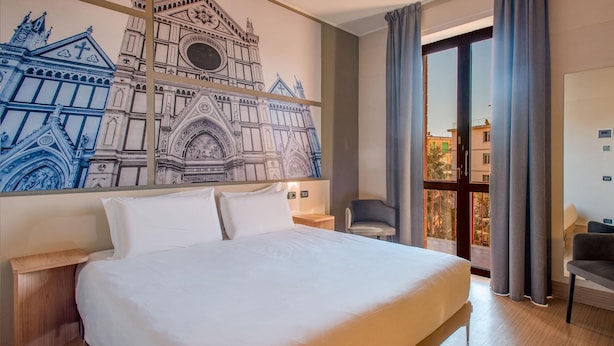 A hotel room with a large bed, a nightstand, a chair, a large window with curtains, and a wall mural of a building.
