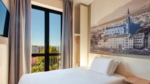 A hotel room with a bed, a TV, a window with curtains, and a cityscape view.