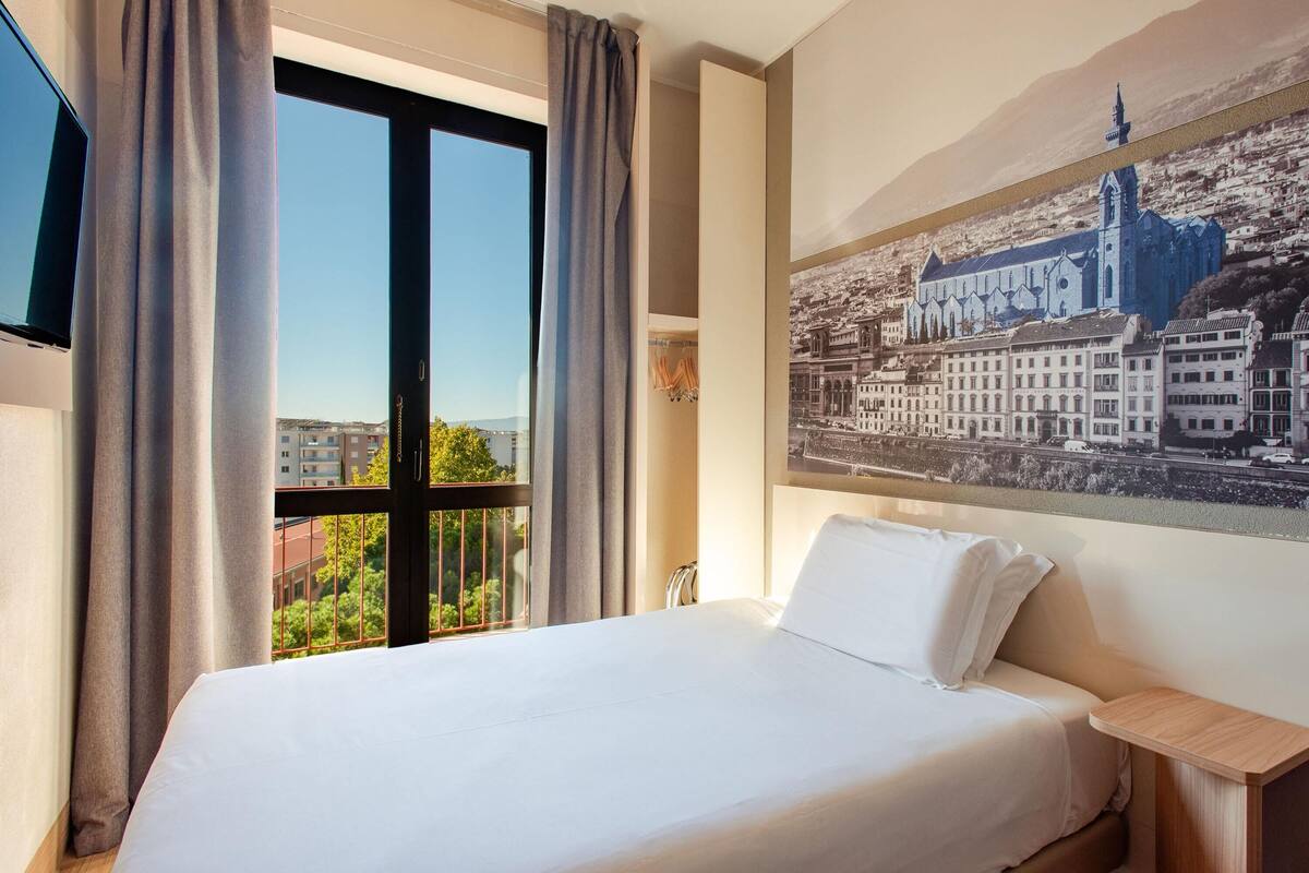 Ein Hotelzimmer mit Bett, Fernseher, Fenster mit Vorhängen und Blick auf die Stadt.