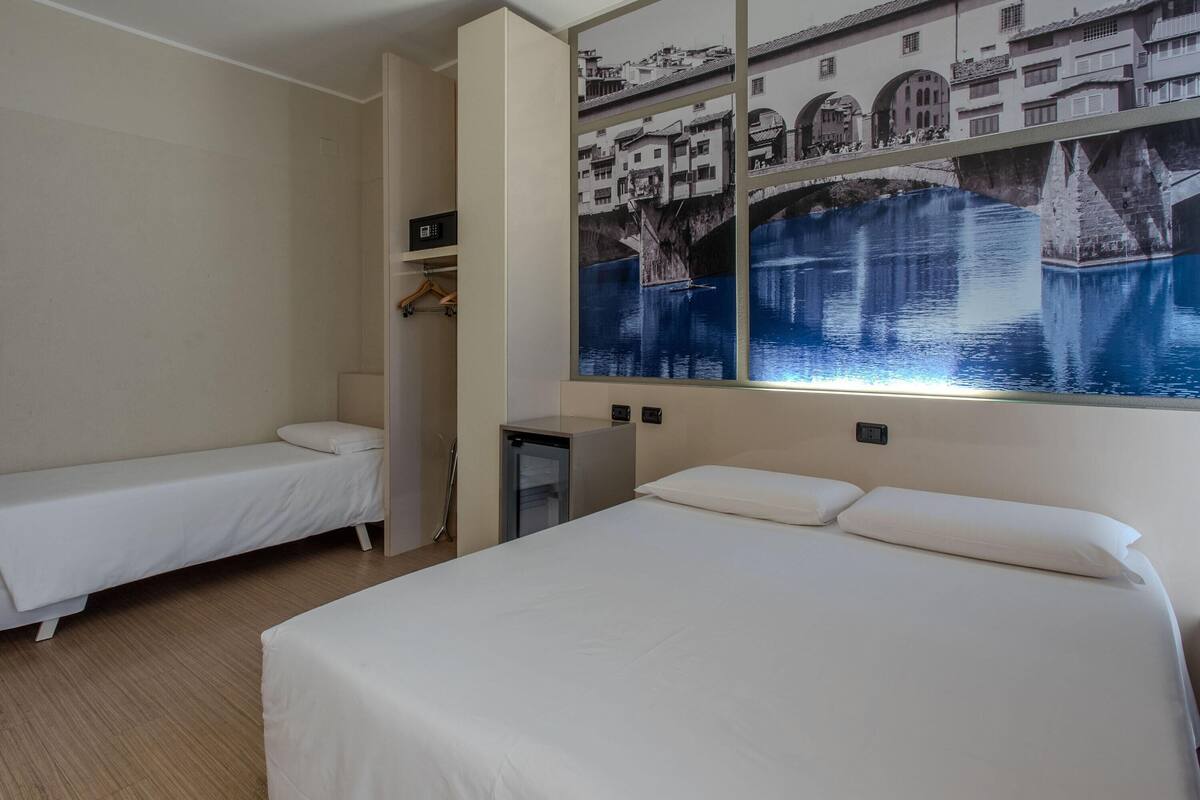 Ein Hotelzimmer mit zwei Einzelbetten, einem kleinen Schreibtisch, einem Kleiderschrank und einem großen Wandbild, das eine Brücke über Wasser zeigt.