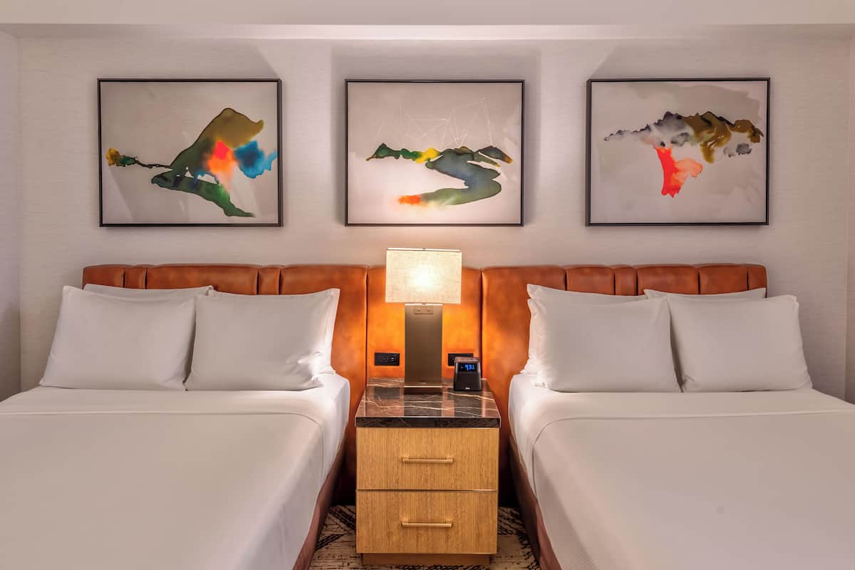 A hotel room with two beds, a nightstand, a lamp, and three abstract wall art pieces.