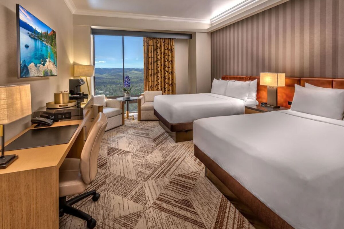 A hotel room with two beds, a desk, a chair, a TV, and a large window with a view.