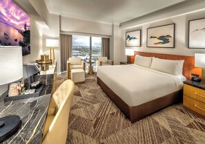 A hotel room with a large bed, a desk with a computer, a chair, a small table with a lamp, and a view of the city.