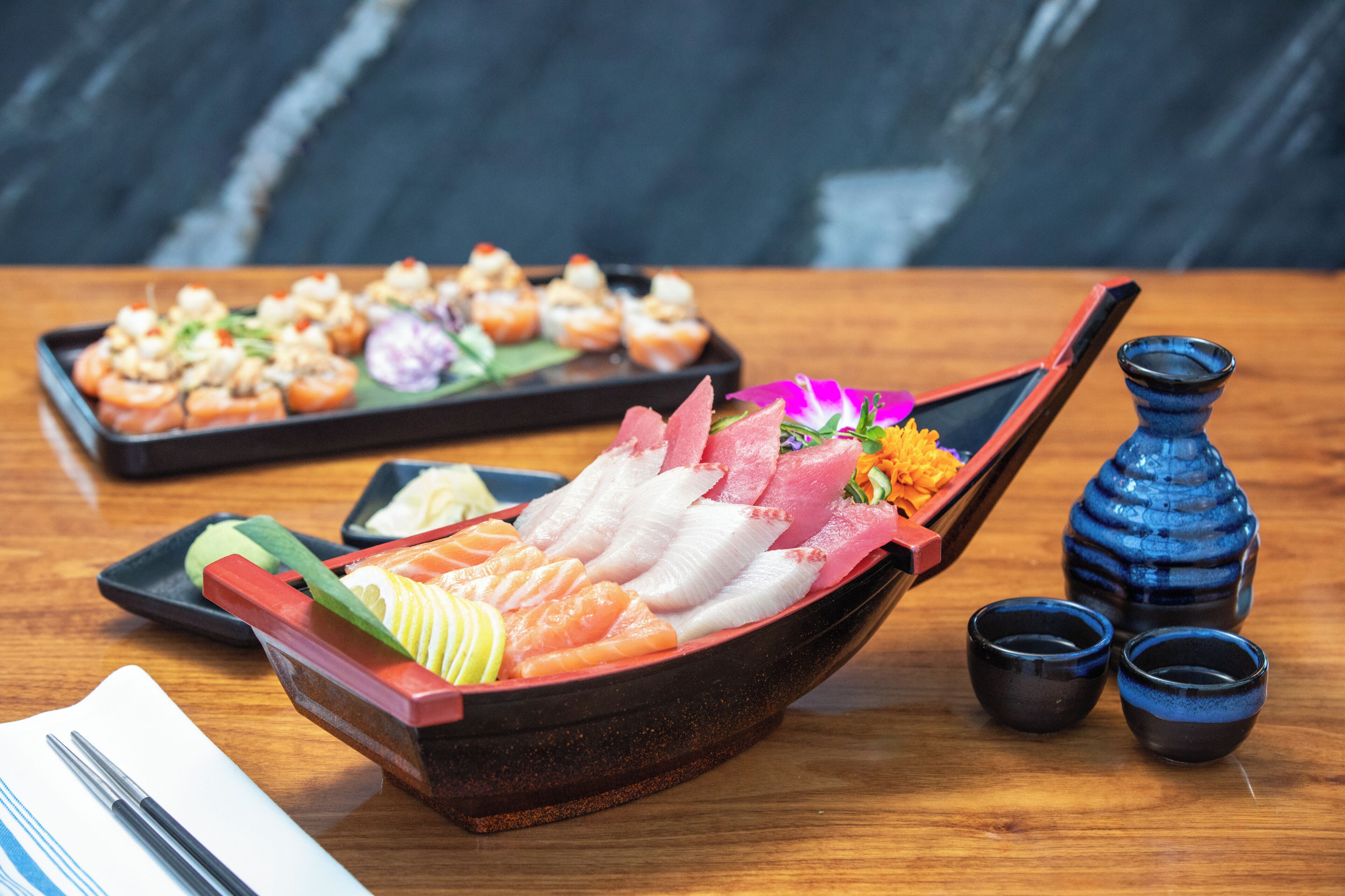 A wooden boat-shaped dish with various types of sushi and sashimi, accompanied by small black cups and a blue vase on a wooden table.