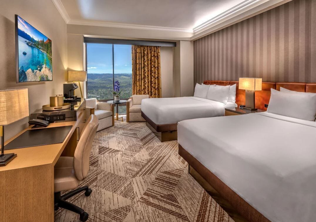 A hotel room with two beds, a desk, a chair, a TV, and a large window with a view.