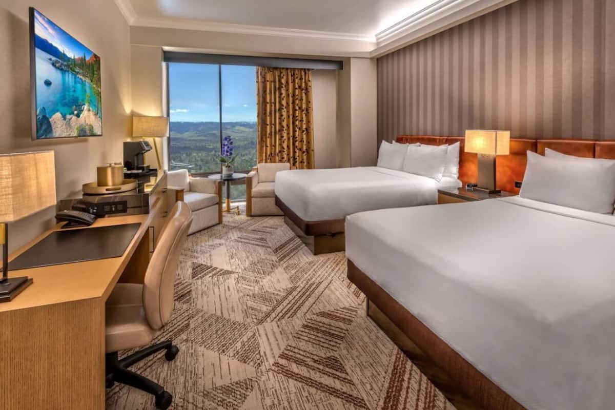 A hotel room with two beds, a desk, a chair, a TV, and a large window with a view.