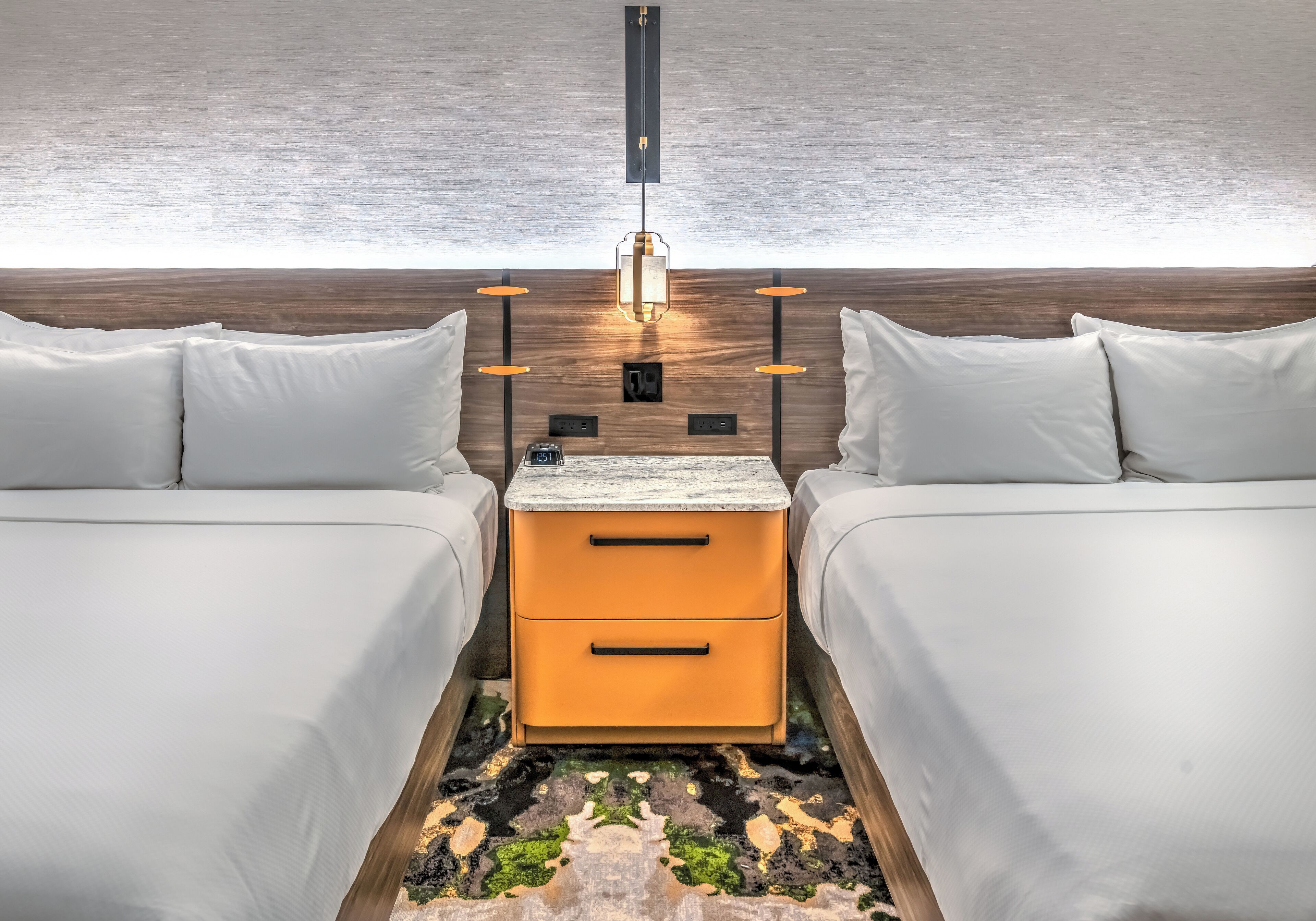A hotel room with two beds, a nightstand, a lamp, and a patterned rug.