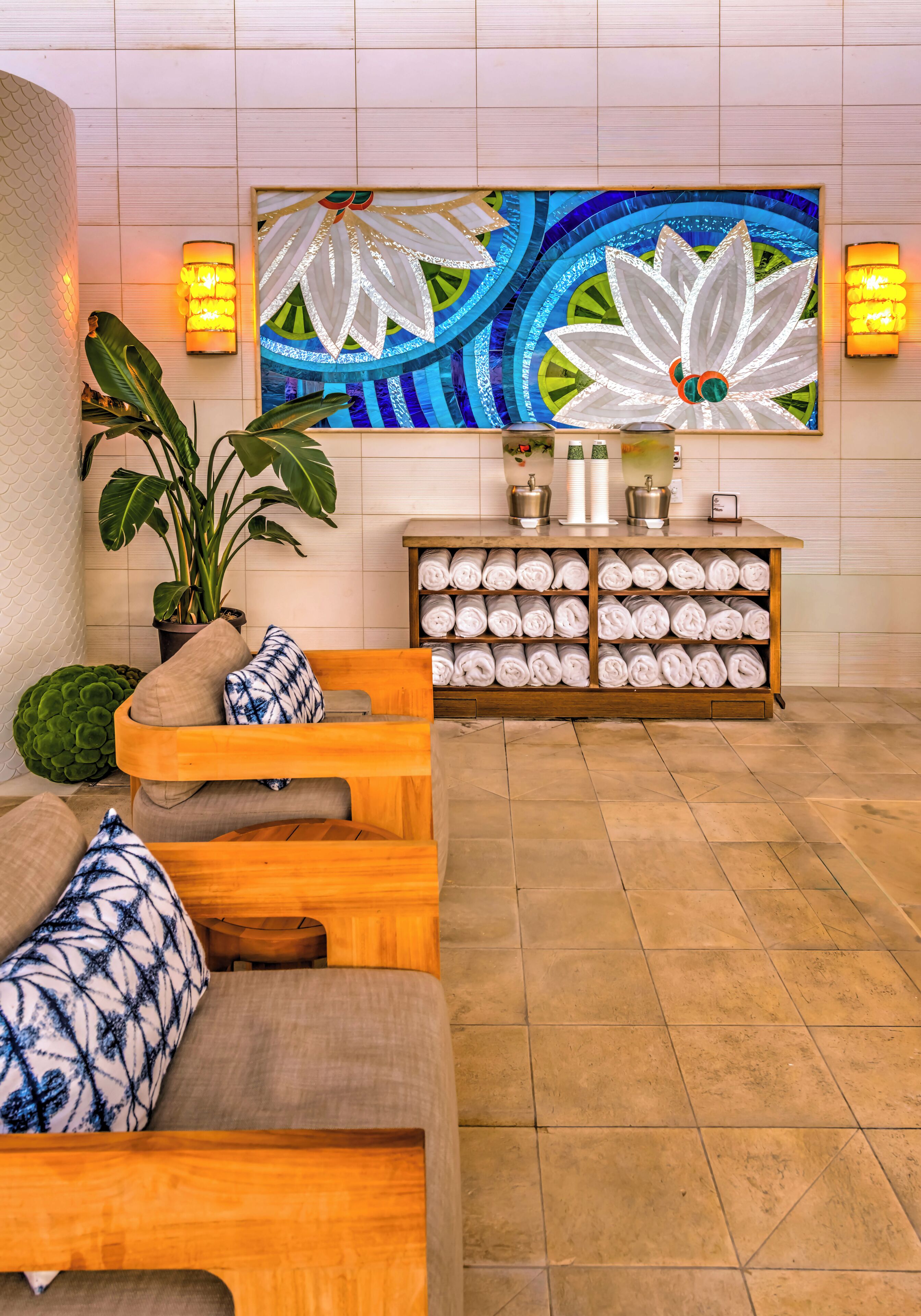 A room with a wooden sofa, a potted plant, a wall-mounted artwork, and a shelf with rolled towels.