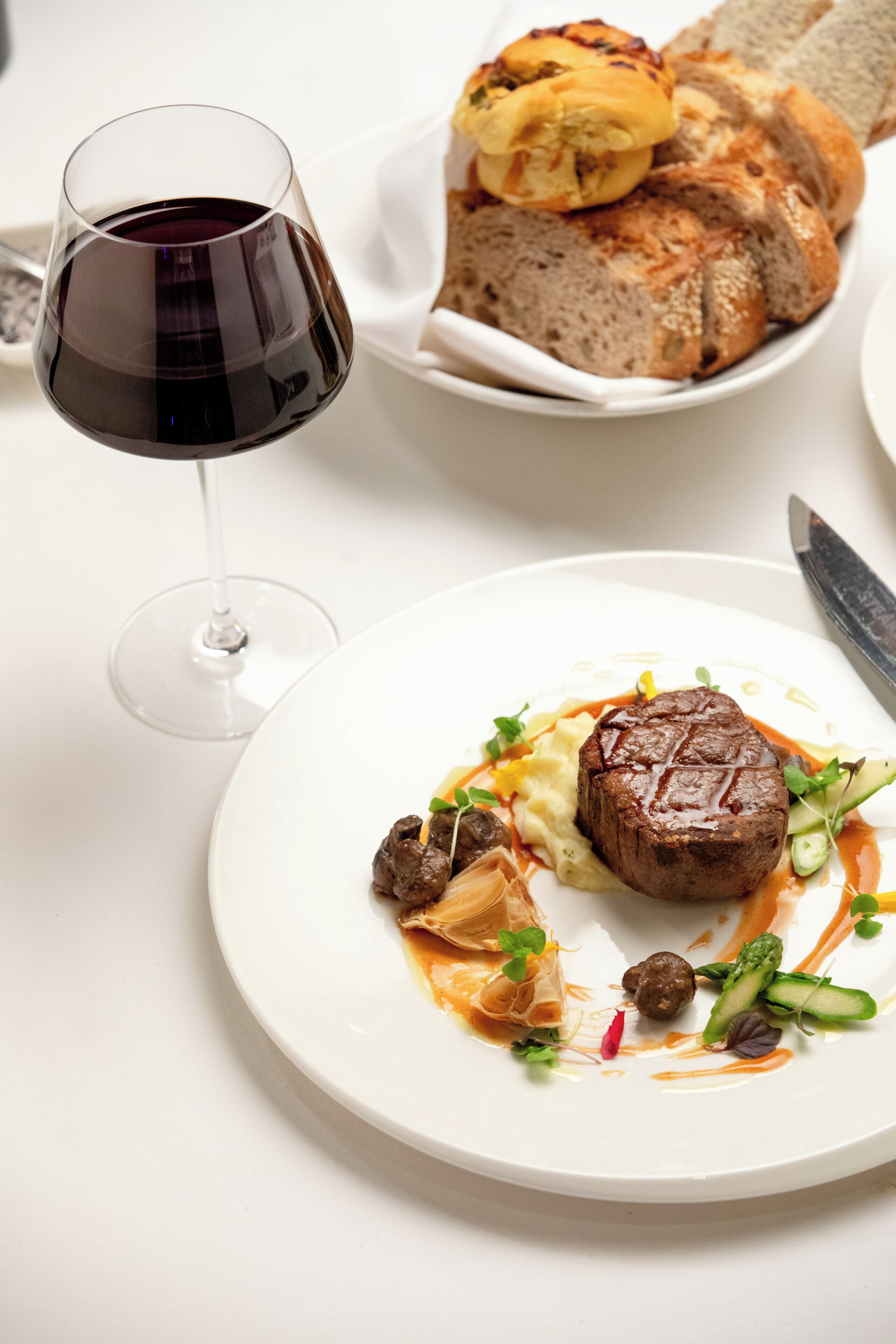 A dining table with a glass of red wine, a plate of food, and a basket of bread.