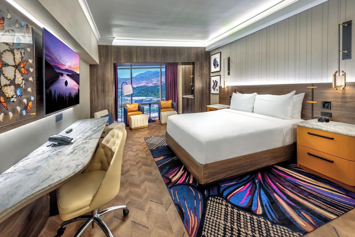 A hotel room with a bed, desk, chair, and a view of mountains through the window.