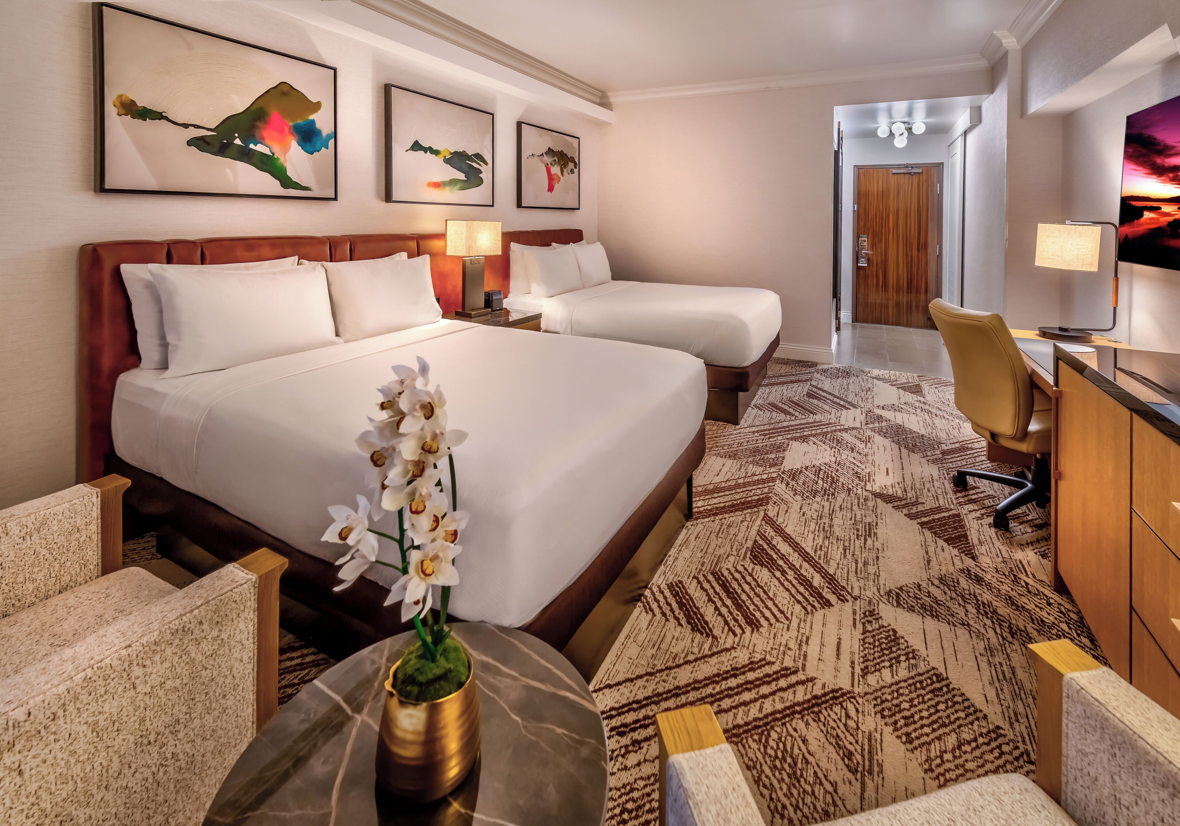 Habitación de hotel con dos camas, un escritorio, una silla y obras de arte en las paredes.