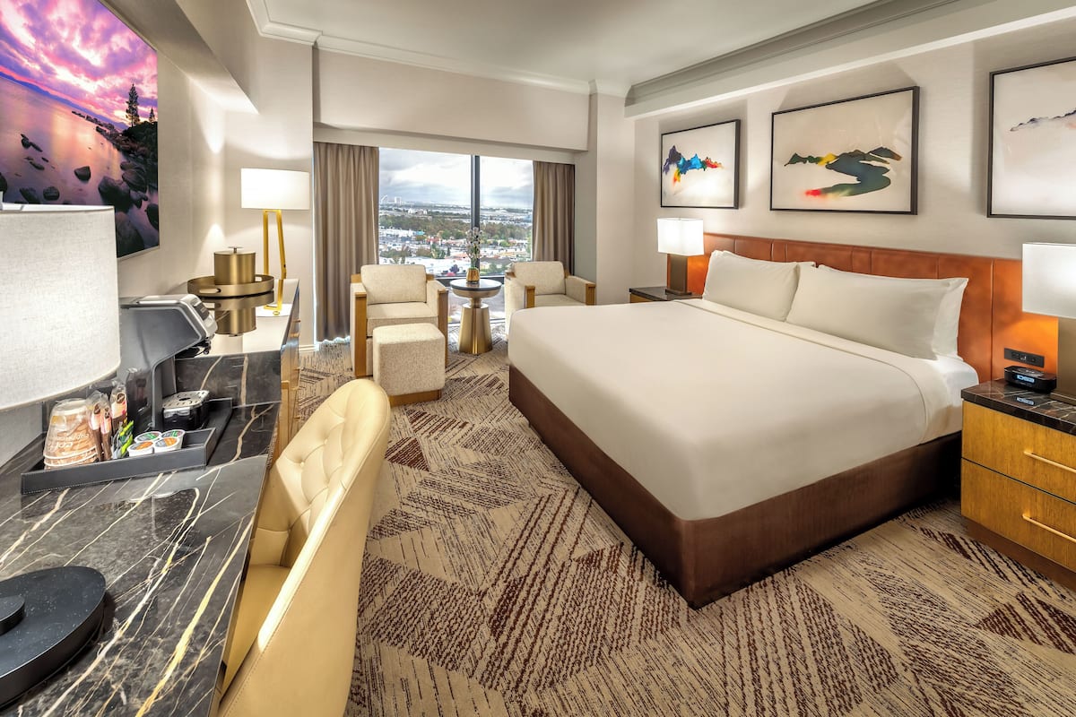 A hotel room with a large bed, a desk with a computer, a chair, a small table with a lamp, and a view of the city.