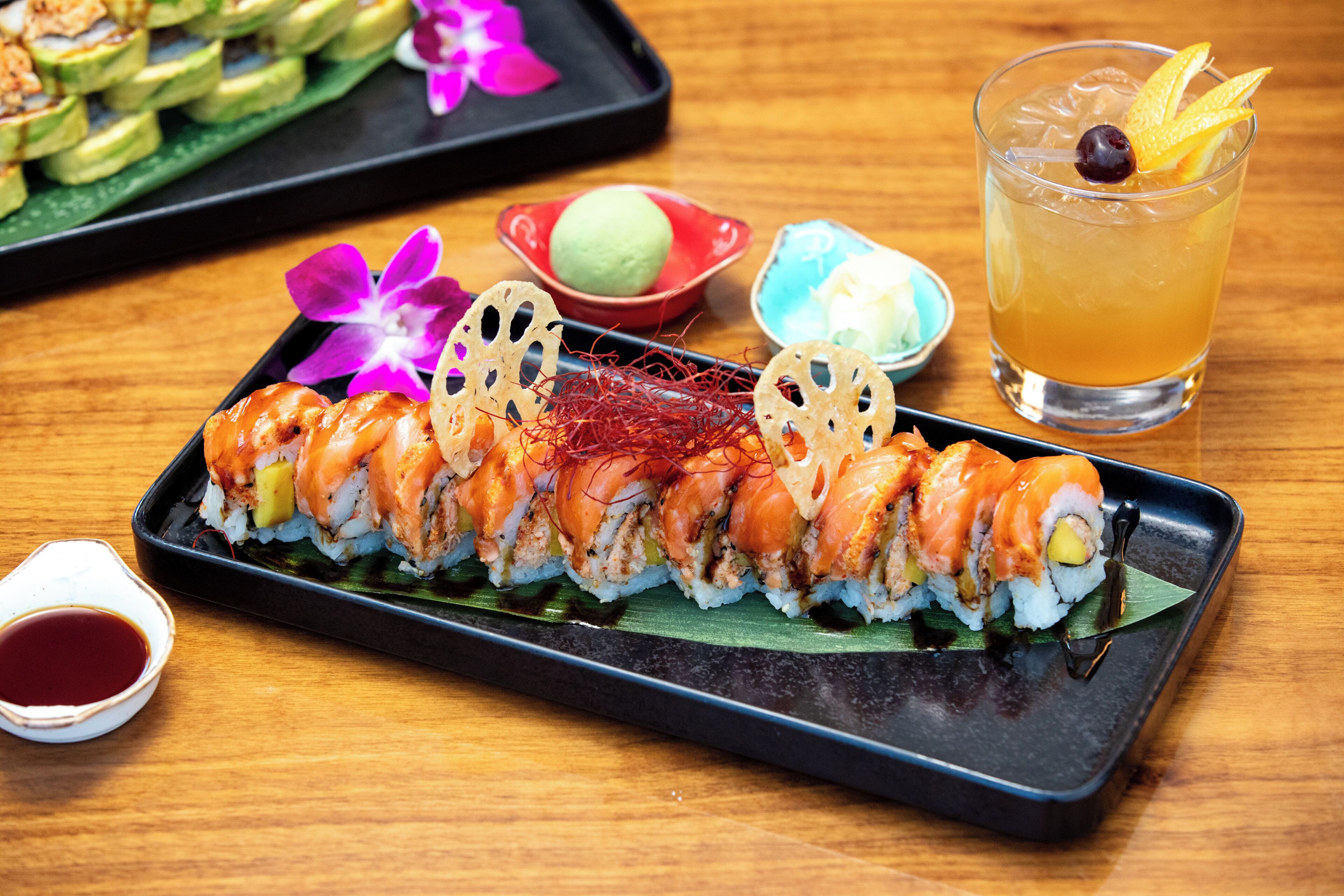 Un plato de sushi con diferentes tipos de sushi, un vaso de té helado y pequeños recipientes con salsas sobre una mesa de madera.