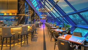 A modern restaurant with a high ceiling and glass walls, featuring a bar area and dining tables.