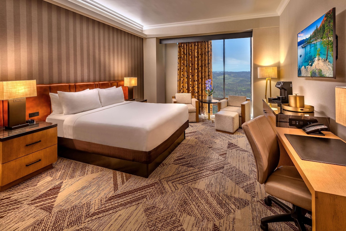 A hotel room with a large bed, a desk with a chair, a small table with chairs, and a large window offering a scenic view.