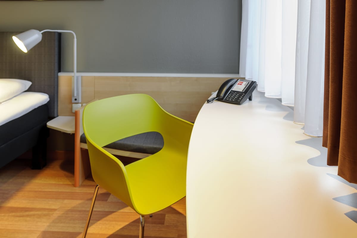 A hotel room with a bed, a desk with a telephone, a bright green chair, and a lamp.