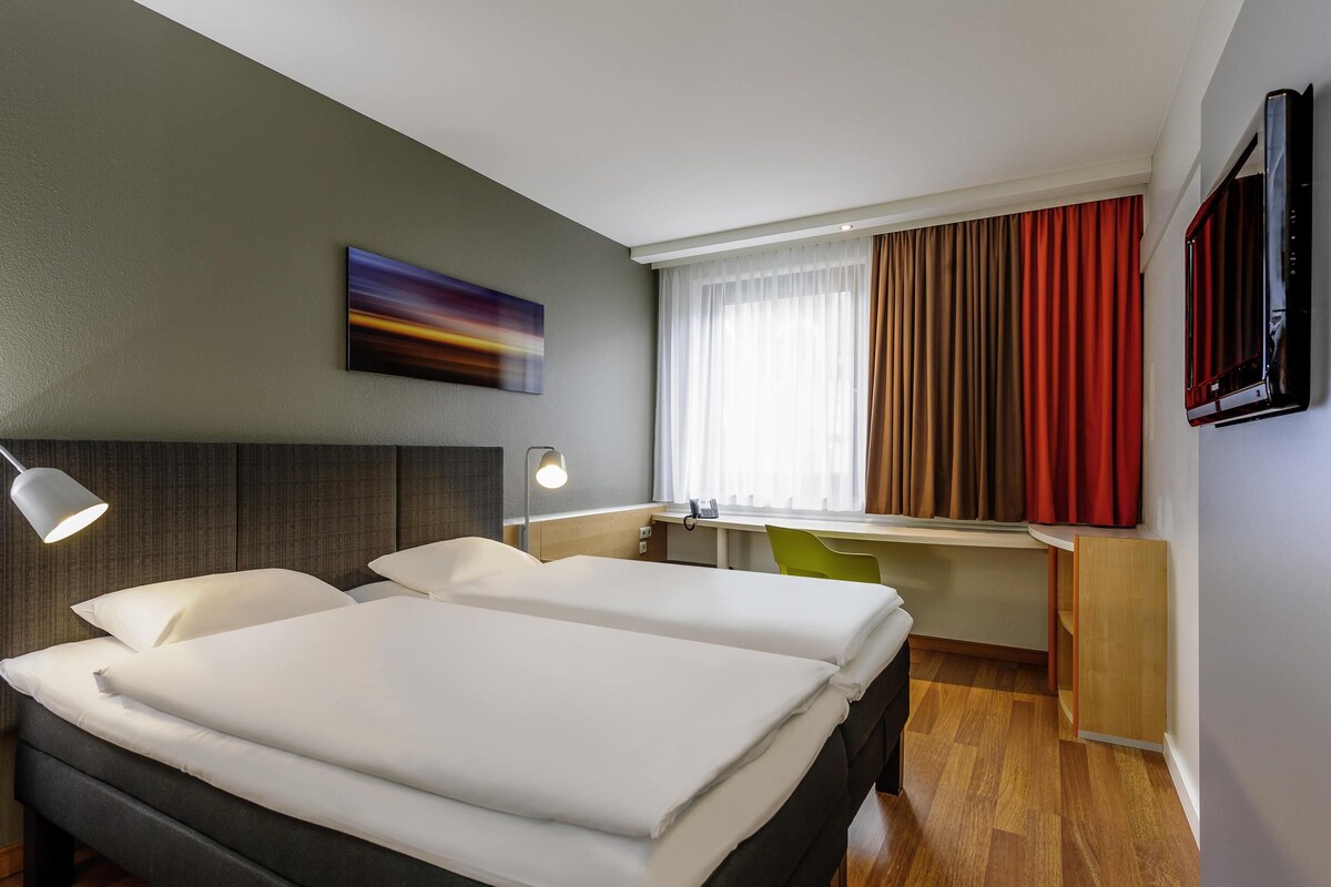 Ein Hotelzimmer mit zwei Betten, einem Schreibtisch, einem Stuhl, einem Fernseher und einem Bild an der Wand.