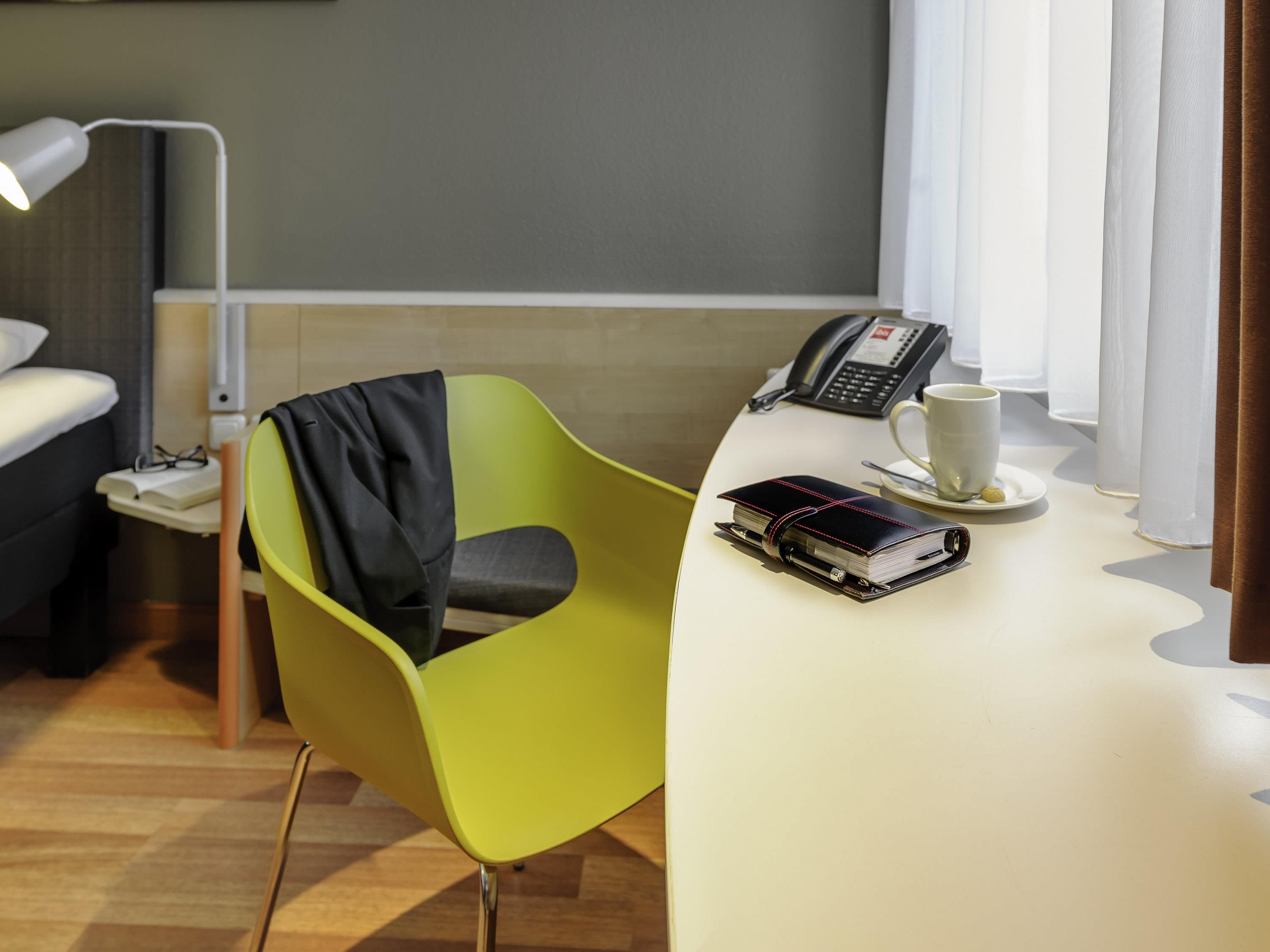 A hotel room with a green chair, a bedside table with a phone, a cup, and a book.