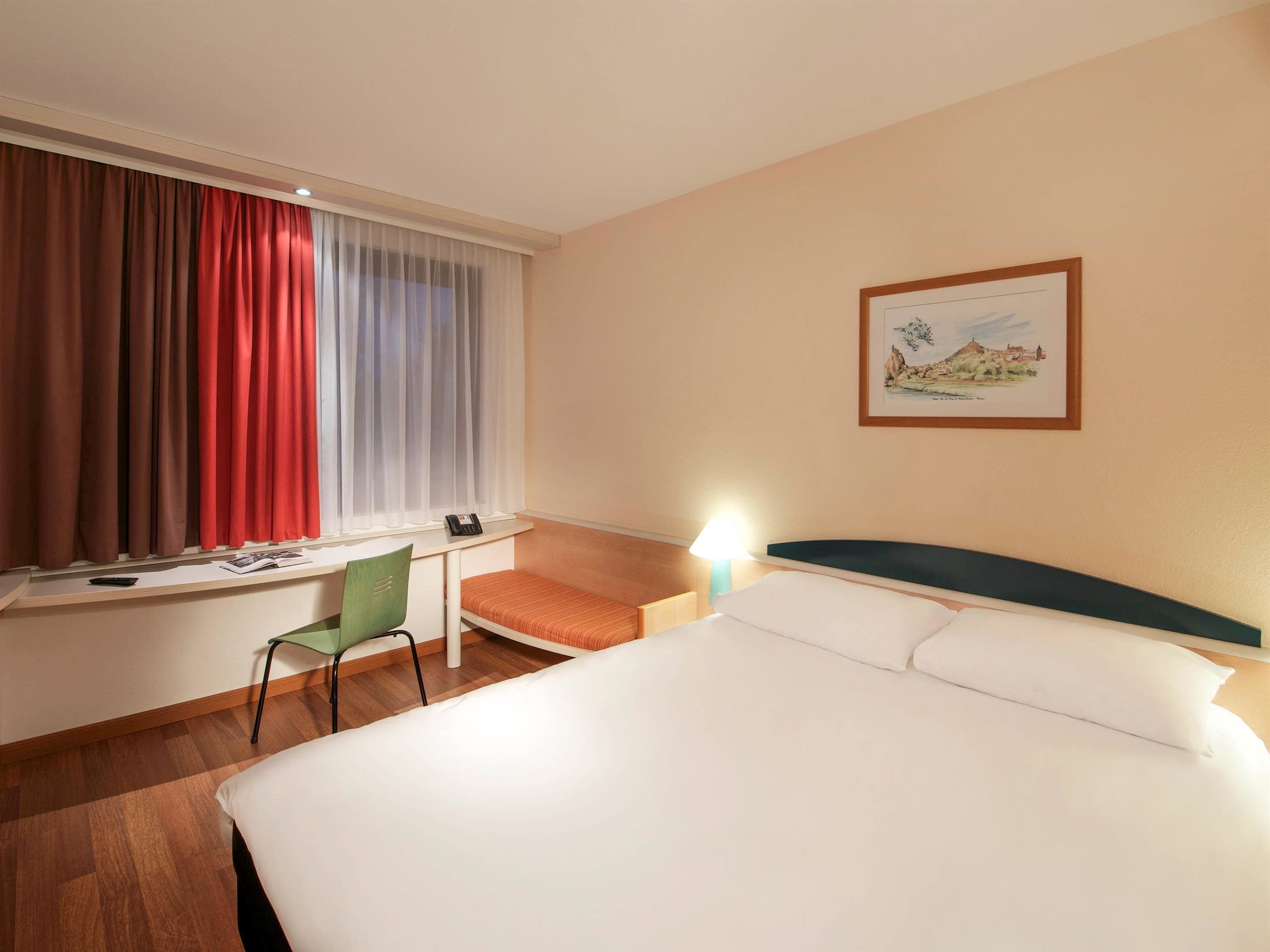 Habitación de hotel con cama, escritorio, silla, ventana con cortinas, lámpara y un cuadro en la pared.