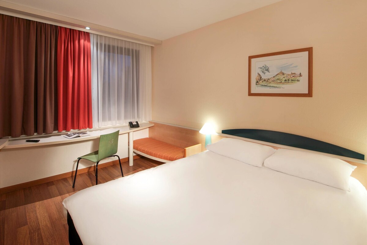 A hotel room with a bed, a desk, a chair, a window with curtains, a lamp, and a framed picture on the wall.