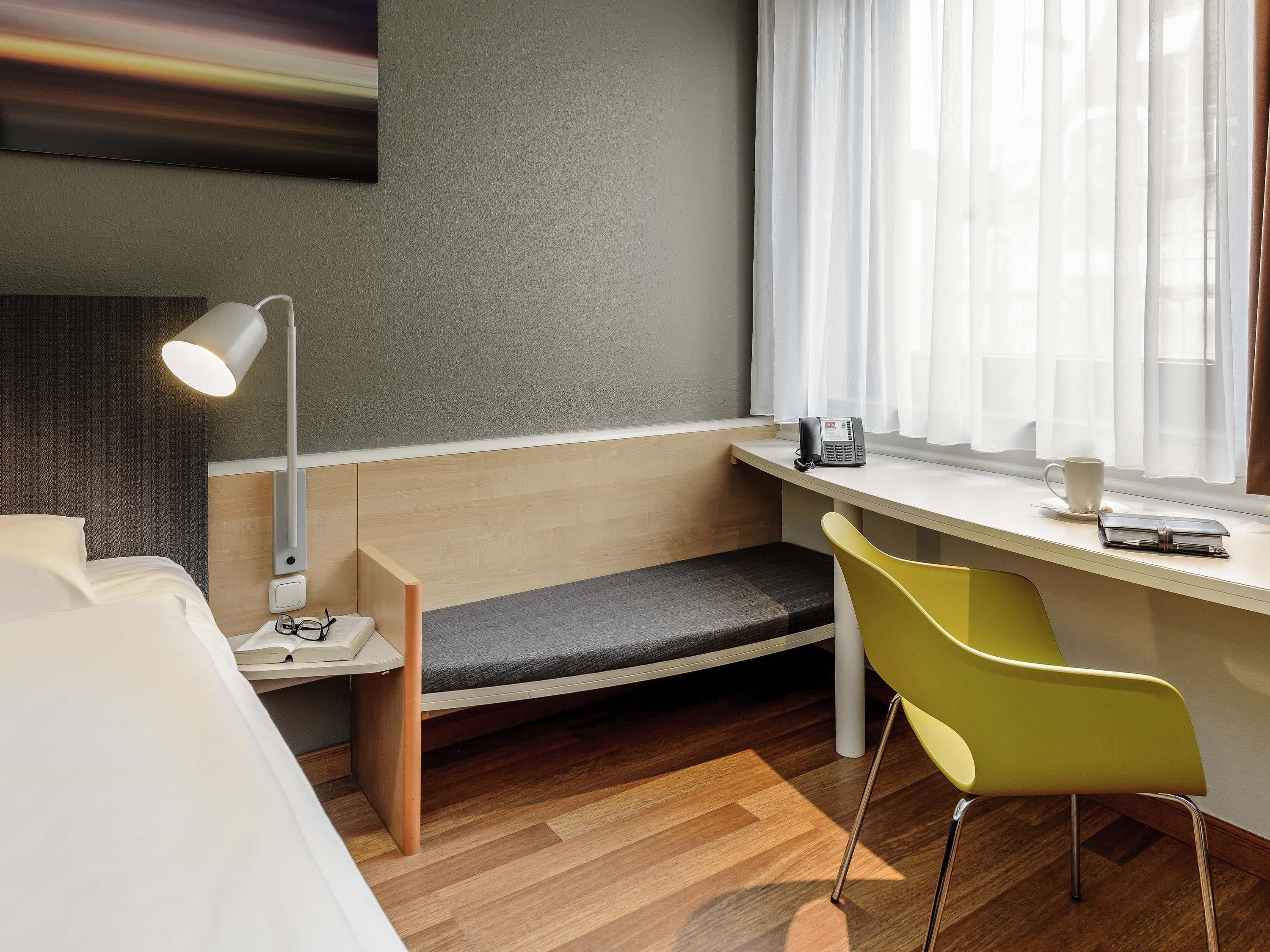 A hotel room with a bed, a desk with a chair, a lamp, a phone, and a window with curtains.