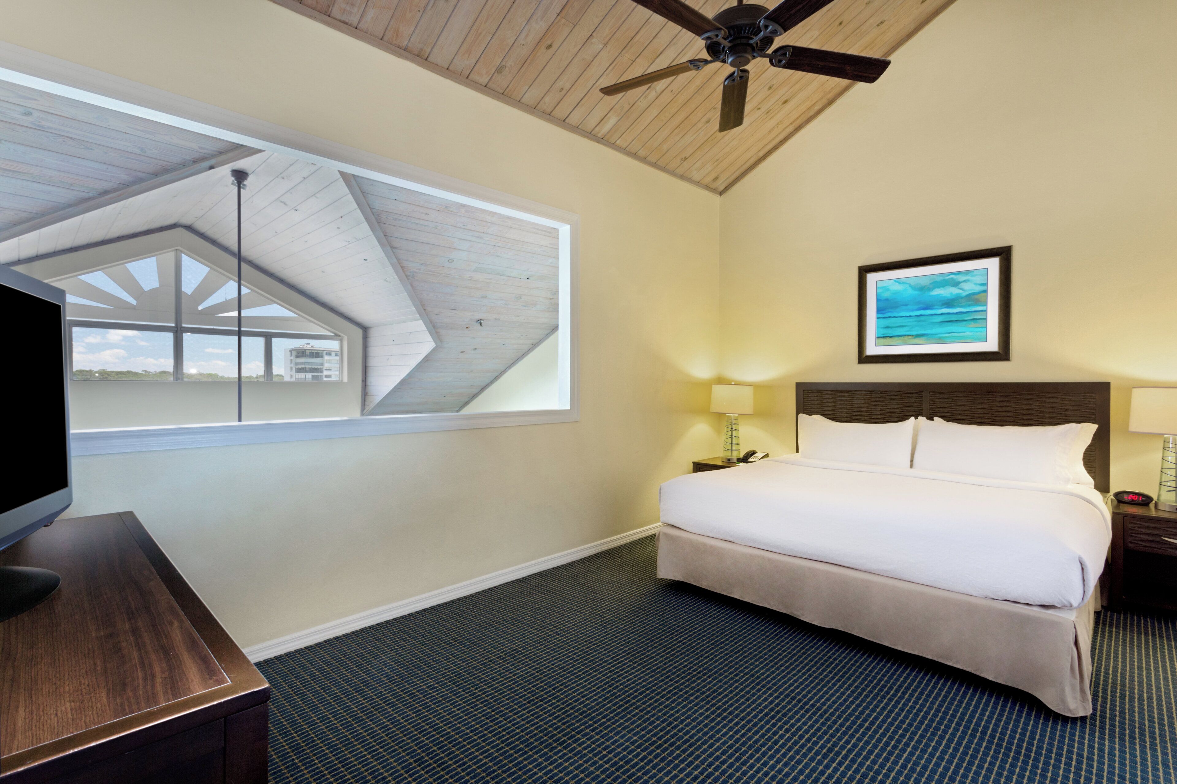 A hotel room with a bed, a desk, a ceiling fan, a window with a view, a framed picture on the wall, and a nightstand with a lamp.