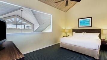 A hotel room with a bed, a desk, a ceiling fan, a window with a view, a framed picture on the wall, and a nightstand with a lamp.