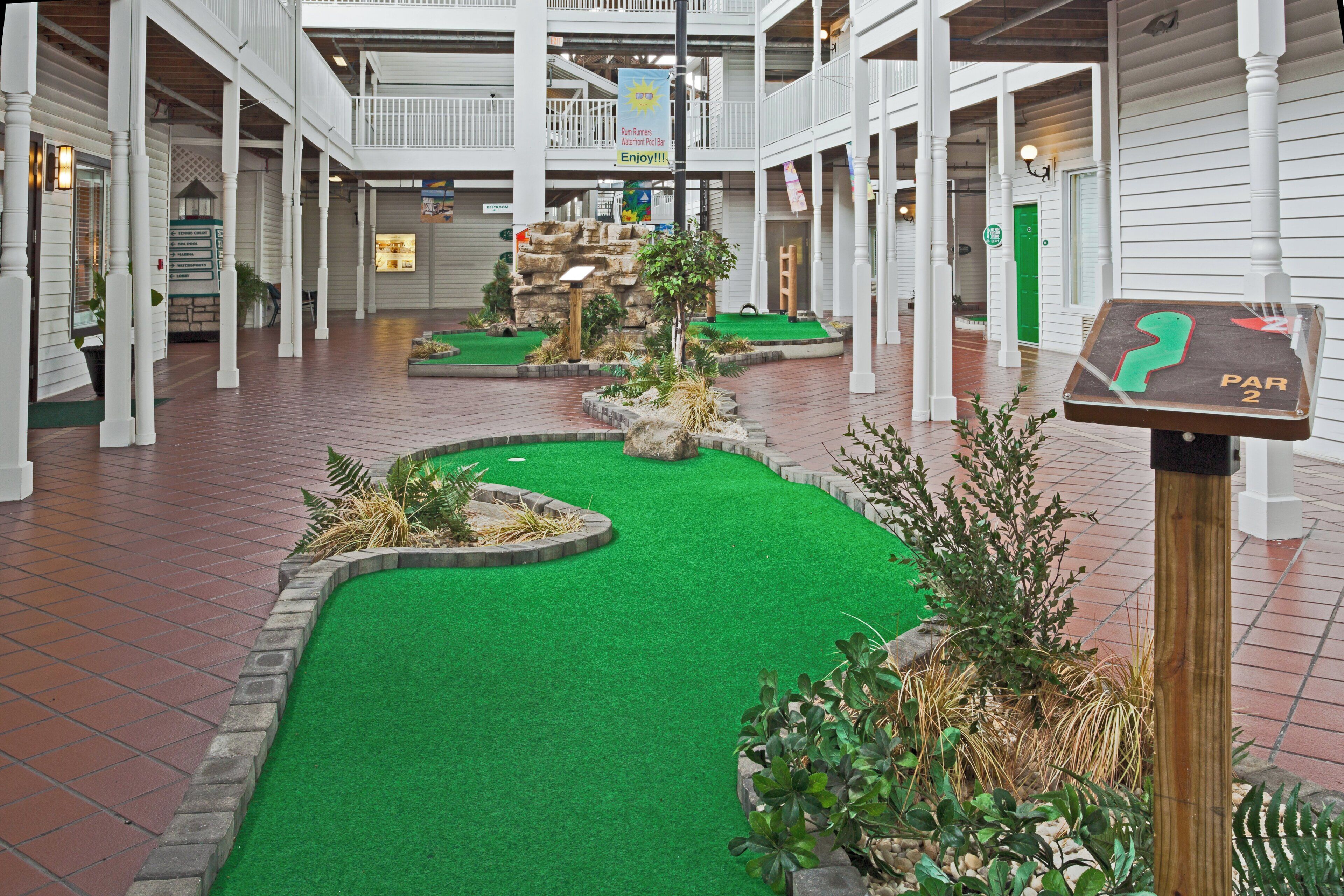Un campo de minigolf con césped artificial y formaciones rocosas, rodeado de pilares blancos y suelo de ladrillo.