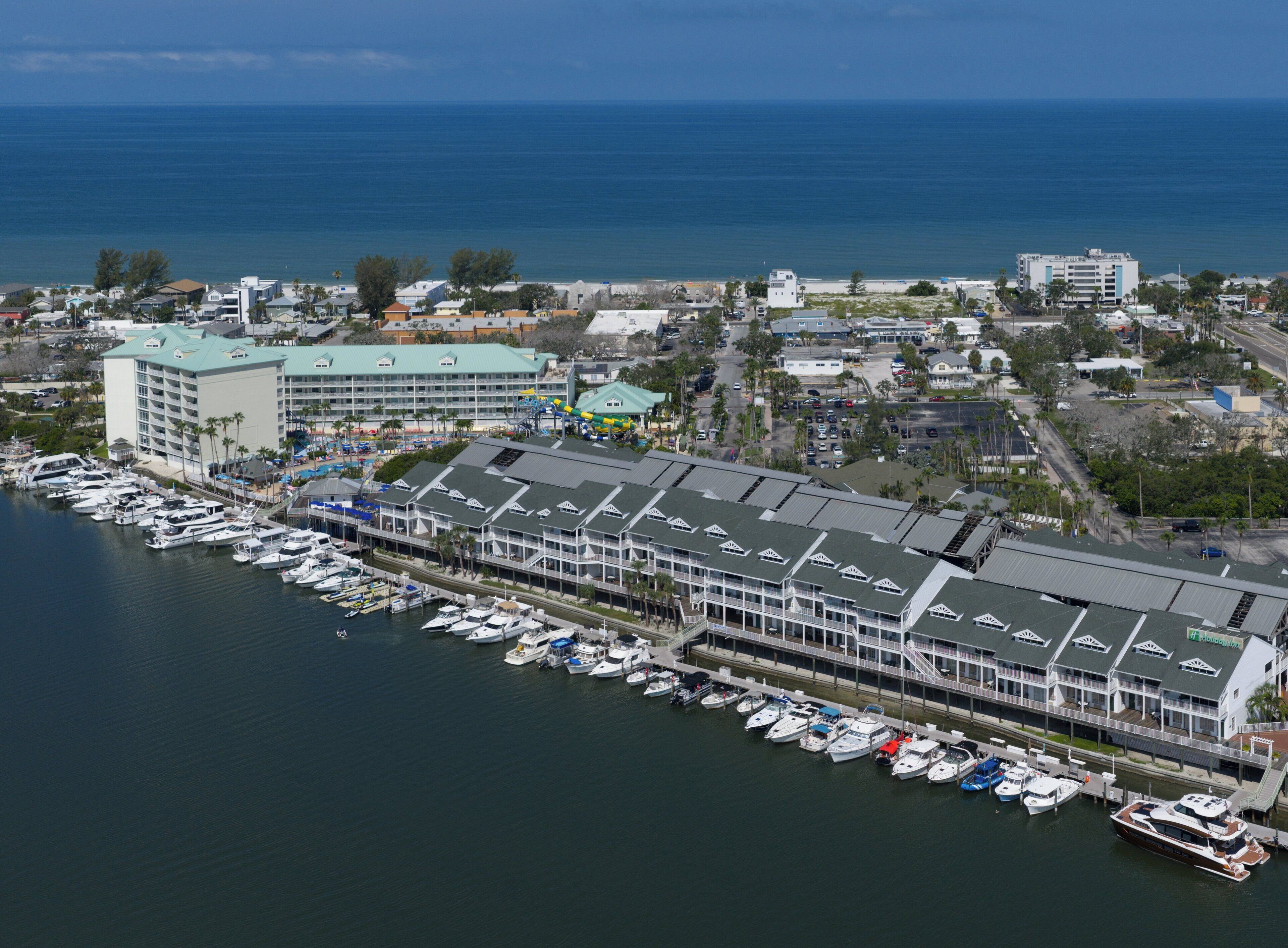 A marina with multiple boats docked, a large hotel with a green roof, and a view of the ocean.
