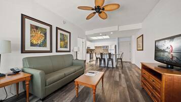 A modern living room with a green sofa, wooden coffee table, and a TV on a wooden cabinet. There is a ceiling fan, framed artwork on the walls, and a kitchen area in the background.