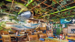 Un restaurante con mesas y sillas de madera, un mural de un barco en la pared y plantas colgantes.