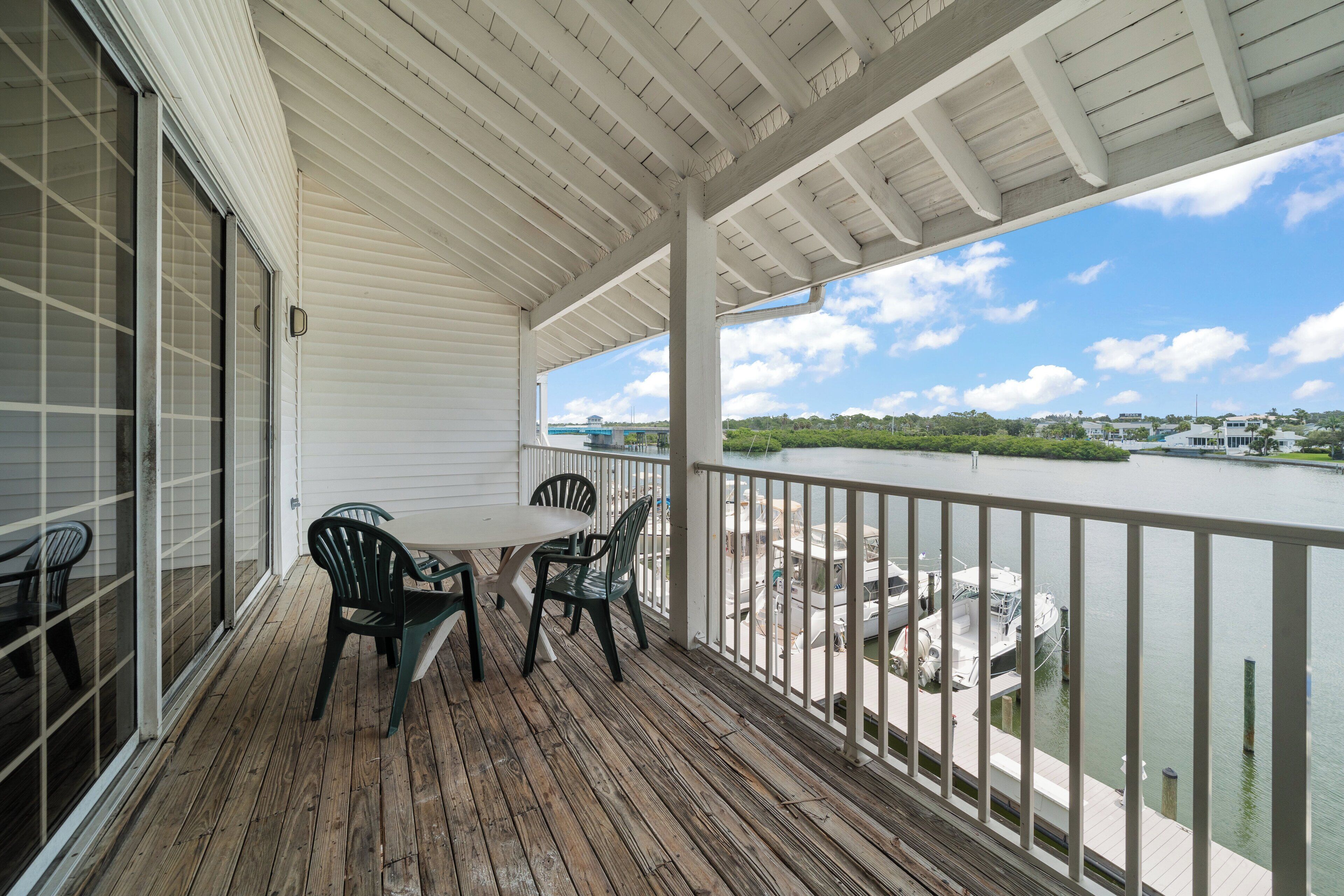Un espace extérieur abrité, doté d’une terrasse en bois, d’une table avec des chaises et offrant une vue sur une rivière et des bâtiments éloignés.