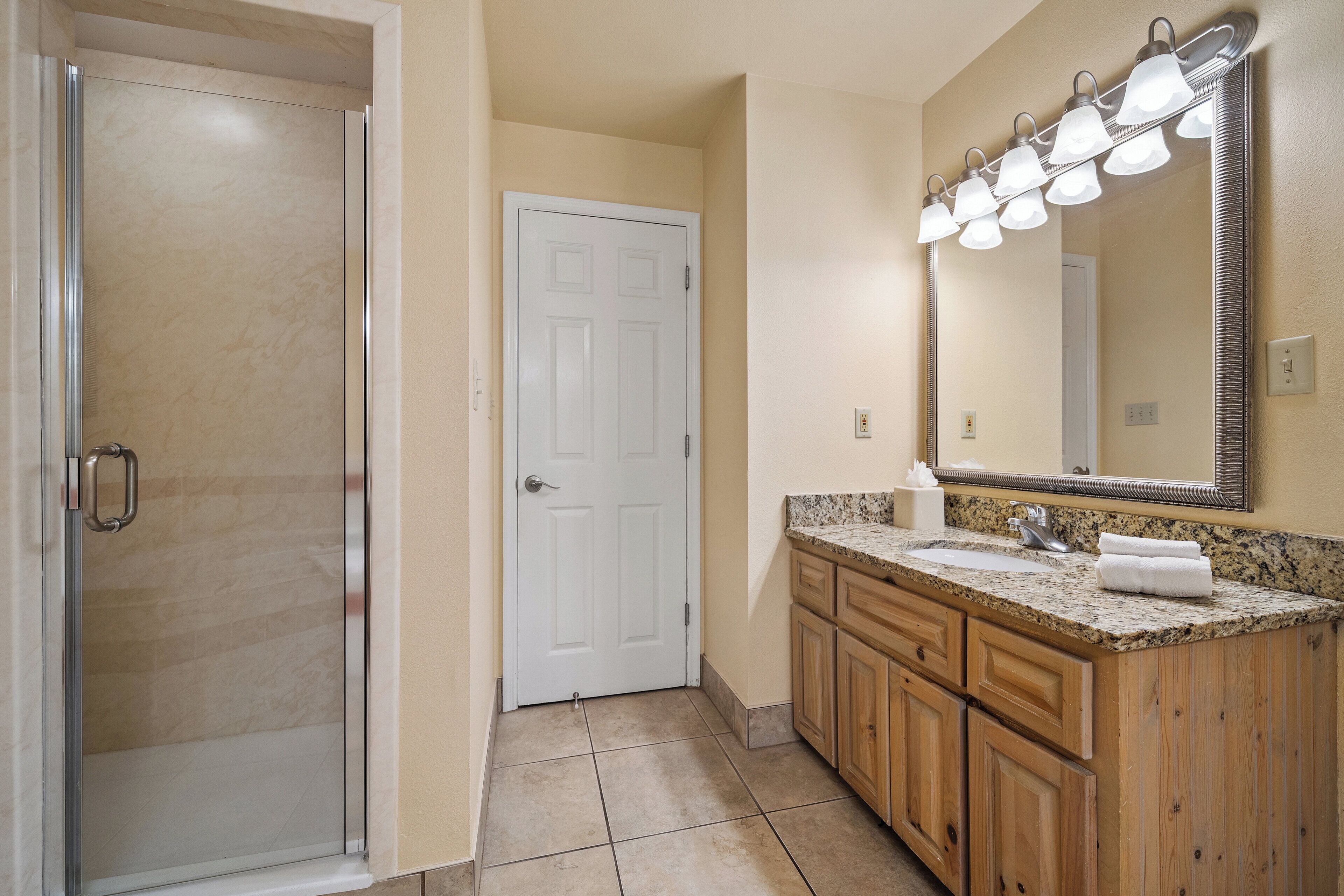 A bathroom with a glass shower enclosure, a white door, a granite countertop sink, wooden cabinets, and a mirror with lights.