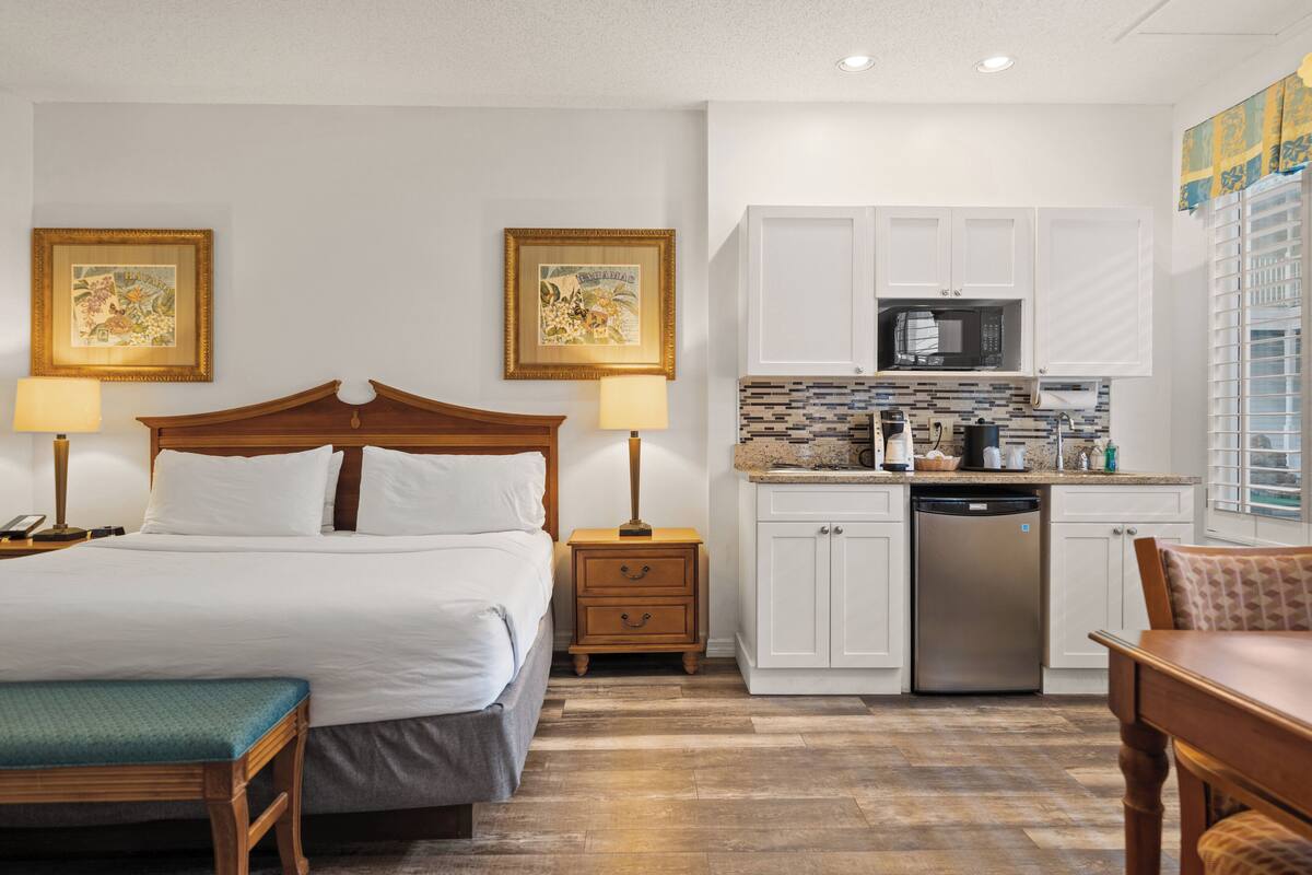 A hotel room with a bed, bedside tables, a desk, a chair, a kitchenette, and a bathroom.