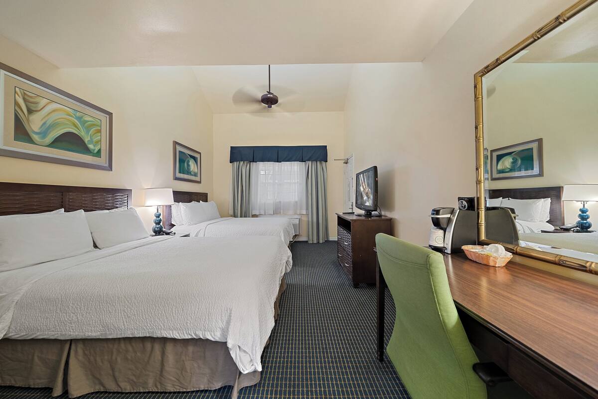 A hotel room with two beds, a desk with a chair, a large mirror, and artwork on the walls.