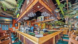 Zona de bar con sillas de madera y mostrador, rodeada de decoración tropical y un mural.
