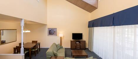 Habitación de hotel con comedor, sofá, mueble para televisión y un amplio ventanal con cortinas translúcidas.