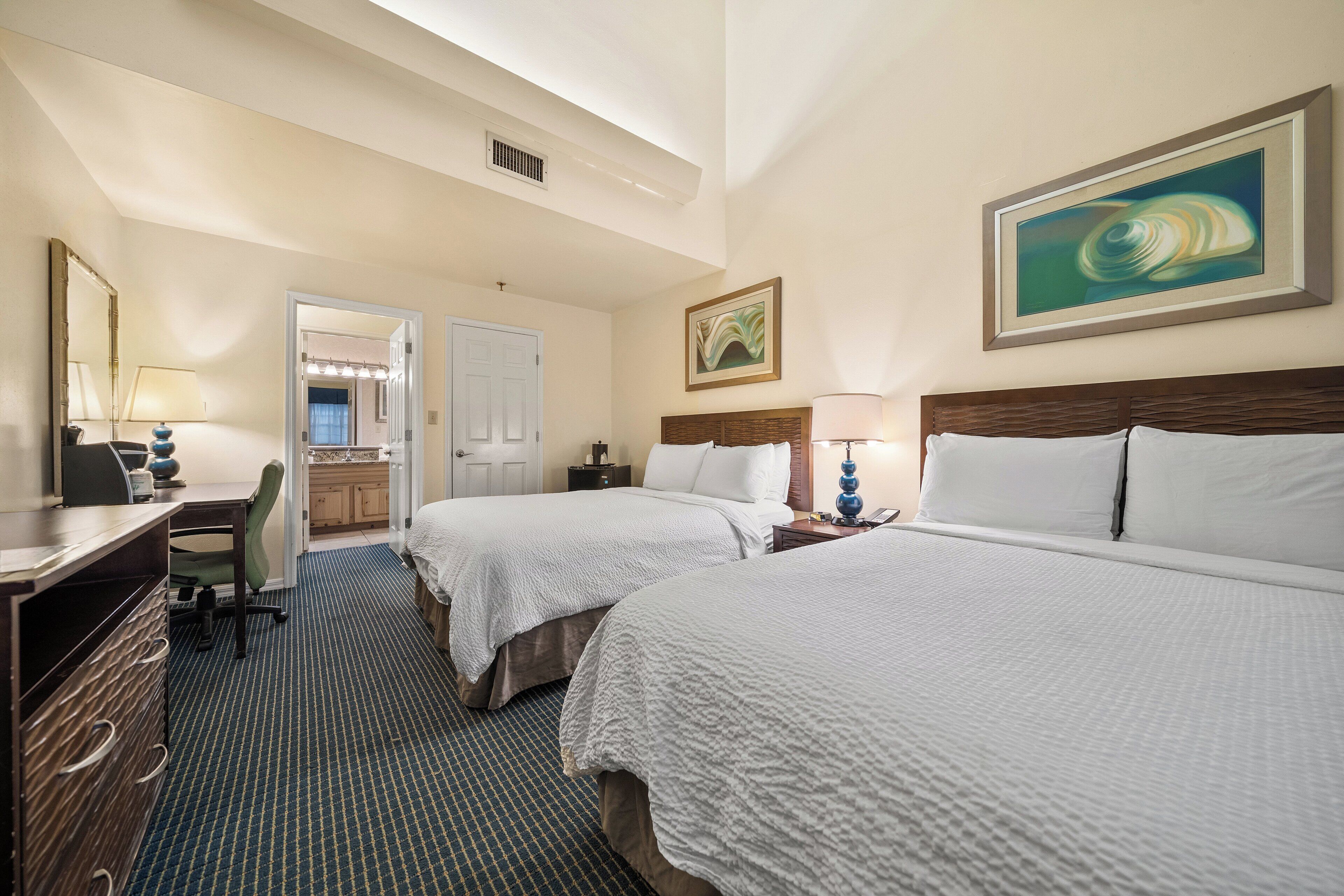 Une chambre d’hôtel avec deux lits, un bureau, une chaise et une salle de bain visible à travers une porte ouverte.