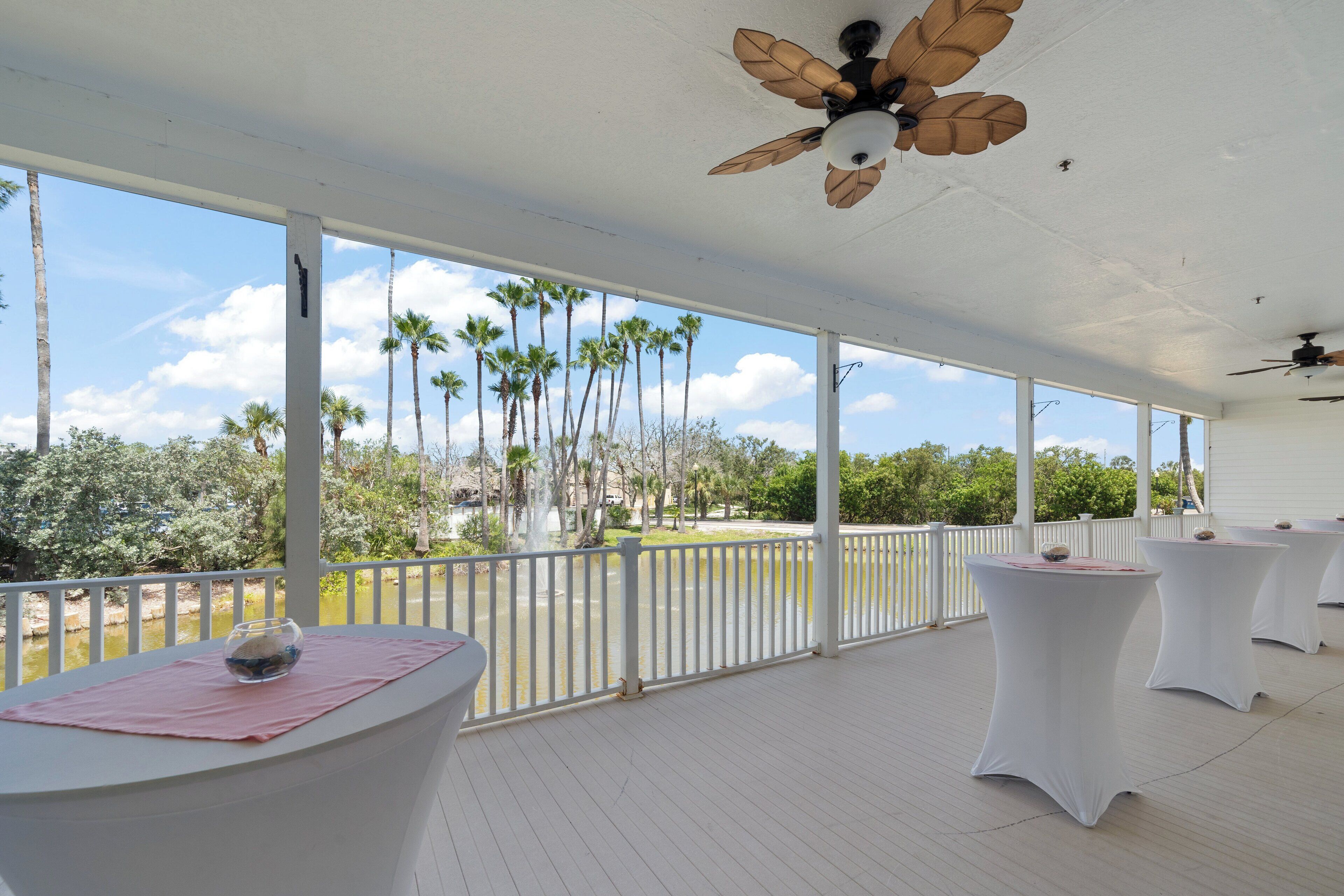 A covered outdoor area with white tables and a view of palm trees and a body of water.