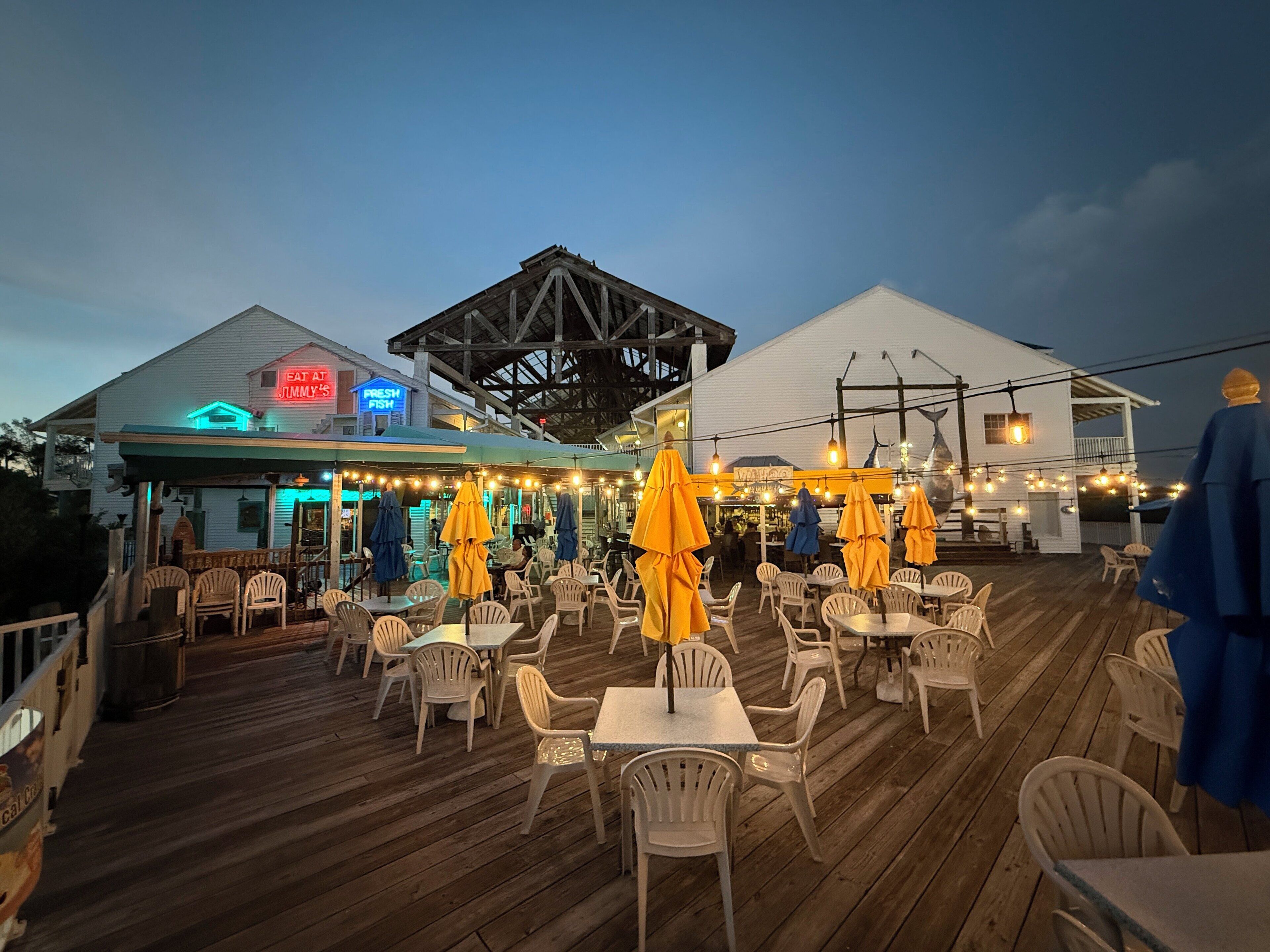 An outdoor dining area with wooden deck and white plastic chairs, featuring a restaurant with neon signs and a covered seating area.