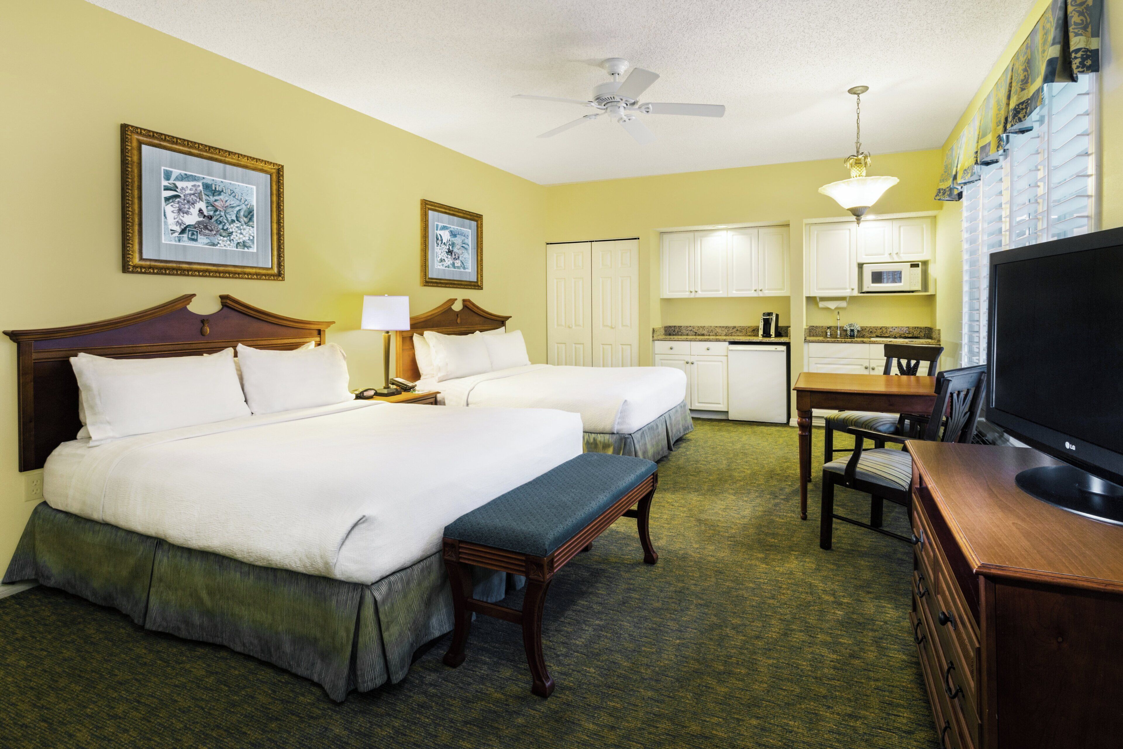 A hotel room with two beds, a desk, a television, and a kitchenette.